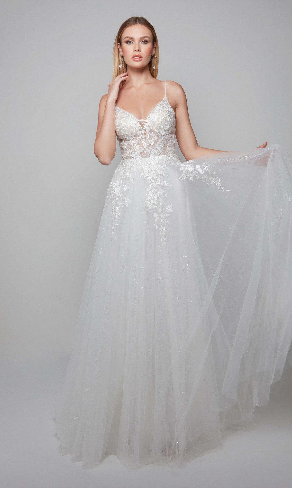 Radiating bridal elegance, she wears the Alyce 7079 long white dress featuring floral lace on the bodice and a sheer tulle skirt, standing against a plain background.